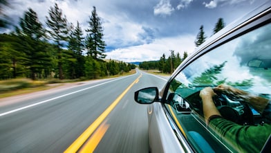Hombre manejando un vehículo por una carretera larga rodeada de árboles.