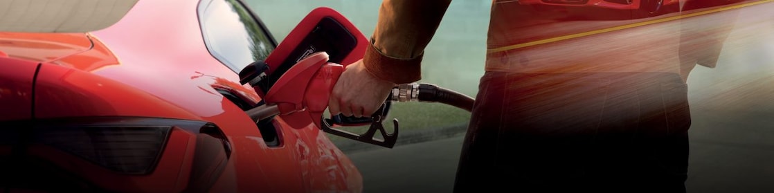 A person refuels a red BMW i4 at a gas station. The car's badge is visible on the rear, and the reflection of another BMW i4 is seen in the person's jacket. The background features an urban skyline with tall buildings and mountains.